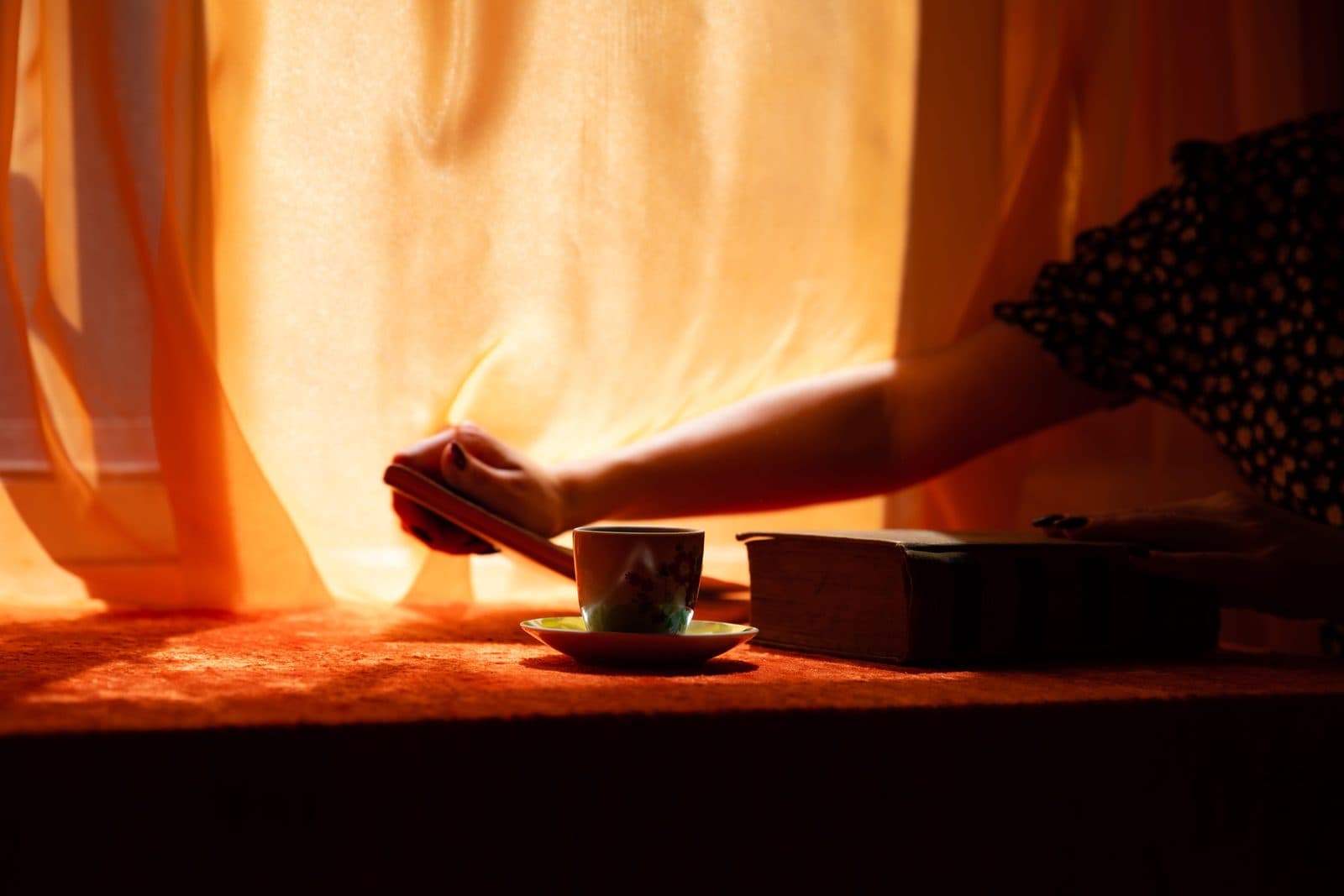 Mão feminina abre cortina junto a xícara de chá e livros em luz âmbar