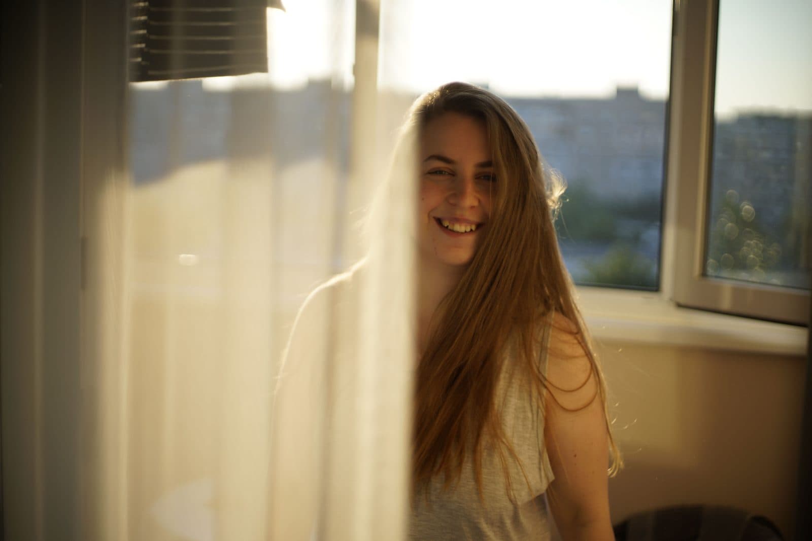 Mulher jovem sorri junto à janela com luz dourada atravessando a cortina